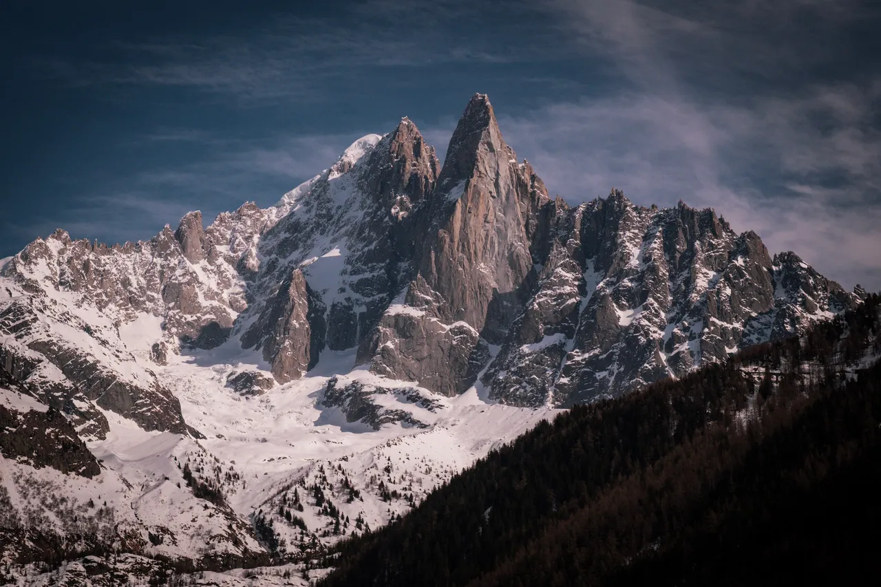 A tall granite spire and a ridge of snowy peaks under a streaked evening sky, above a dark forested slope.