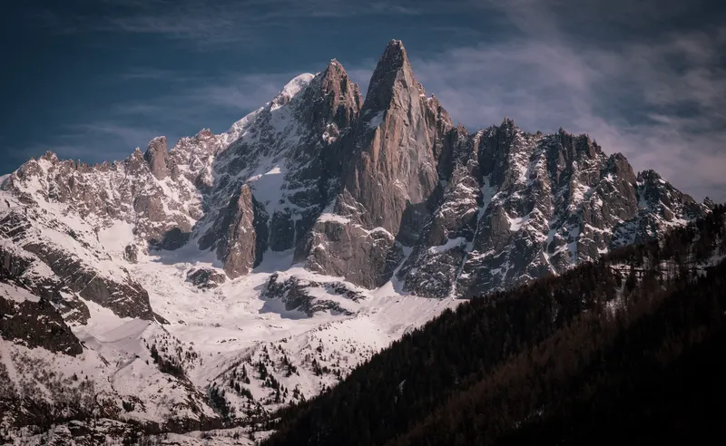 A tall granite spire and a ridge of snowy peaks under a streaked evening sky, above a dark forested slope.