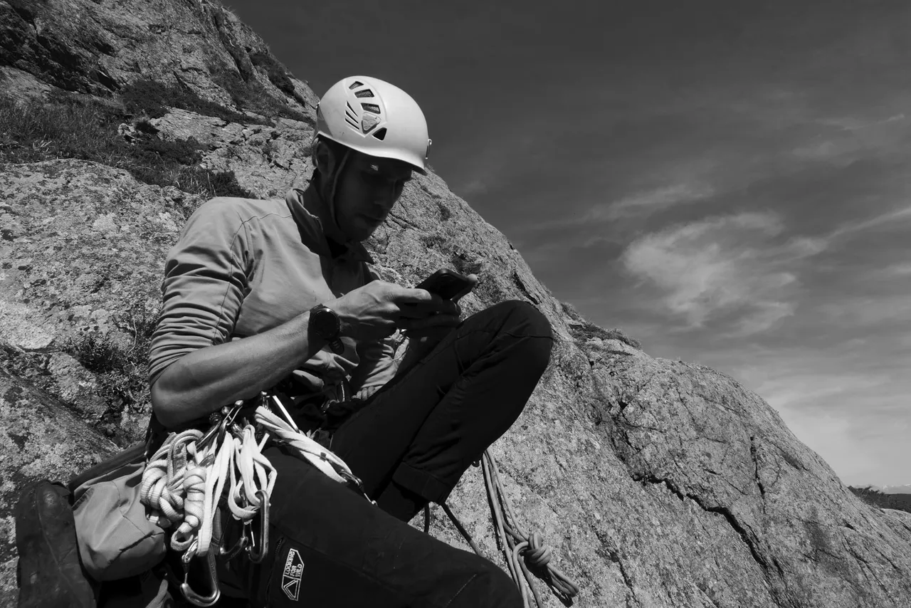 A climber in a white helmet sitting on a sloping granite slab with gear on their harness, looking at a phone. Black and white.