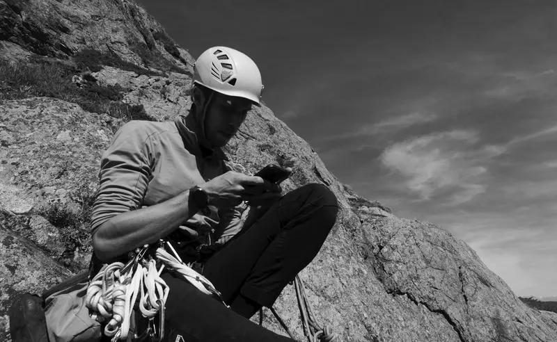 A climber in a white helmet sitting on a sloping granite slab with gear on their harness, looking at a phone. Black and white.