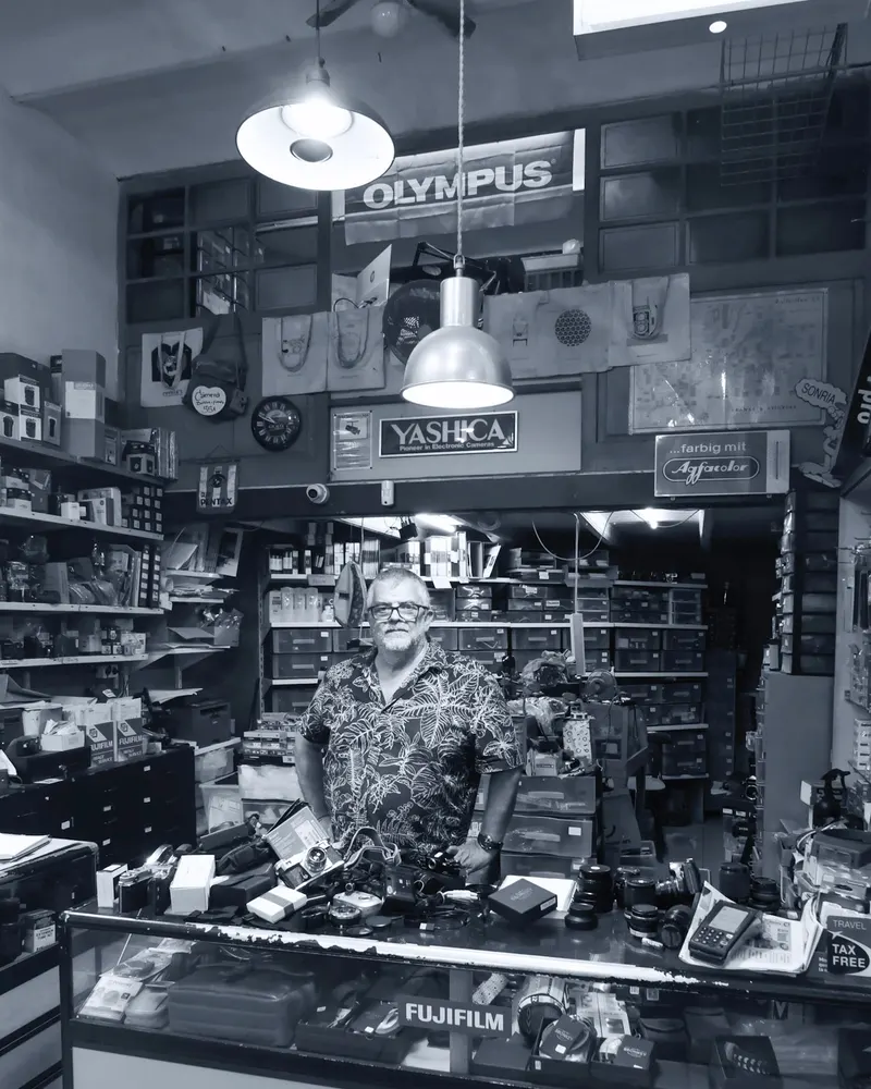 A man in a patterned shirt standing behind the counter of a crowded camera shop. Black and white.