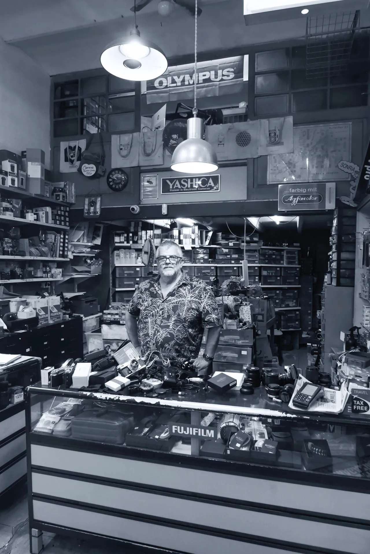 A man in a patterned shirt standing behind the counter of a crowded camera shop. Black and white.