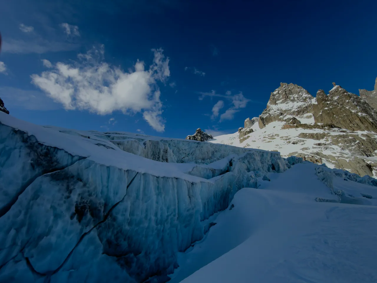 A wall of blue glacier ice beside a crevasse, with a rocky ridge beyond under a deep blue sky.