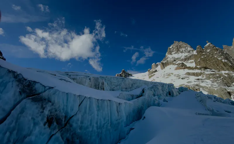 A wall of blue glacier ice beside a crevasse, with a rocky ridge beyond under a deep blue sky.