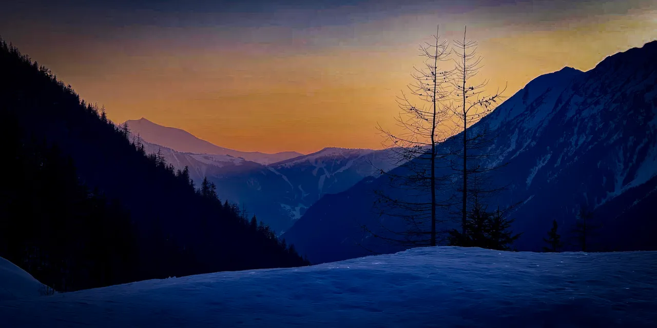 Dusk over a snowy valley, with bare trees and layered mountains against an orange sky.