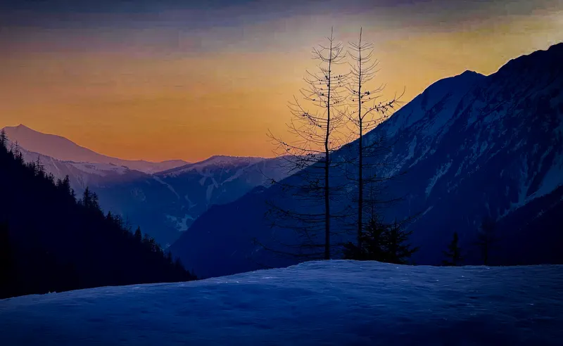 Dusk over a snowy valley, with bare trees and layered mountains against an orange sky.