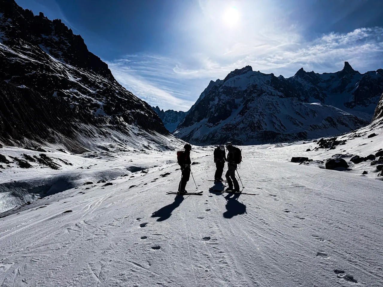 Three ski tourers standing on a wide glacier, backlit by the sun, between dark rock walls.
