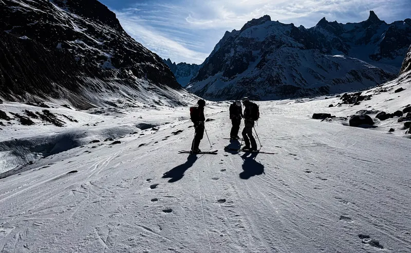 Three ski tourers standing on a wide glacier, backlit by the sun, between dark rock walls.