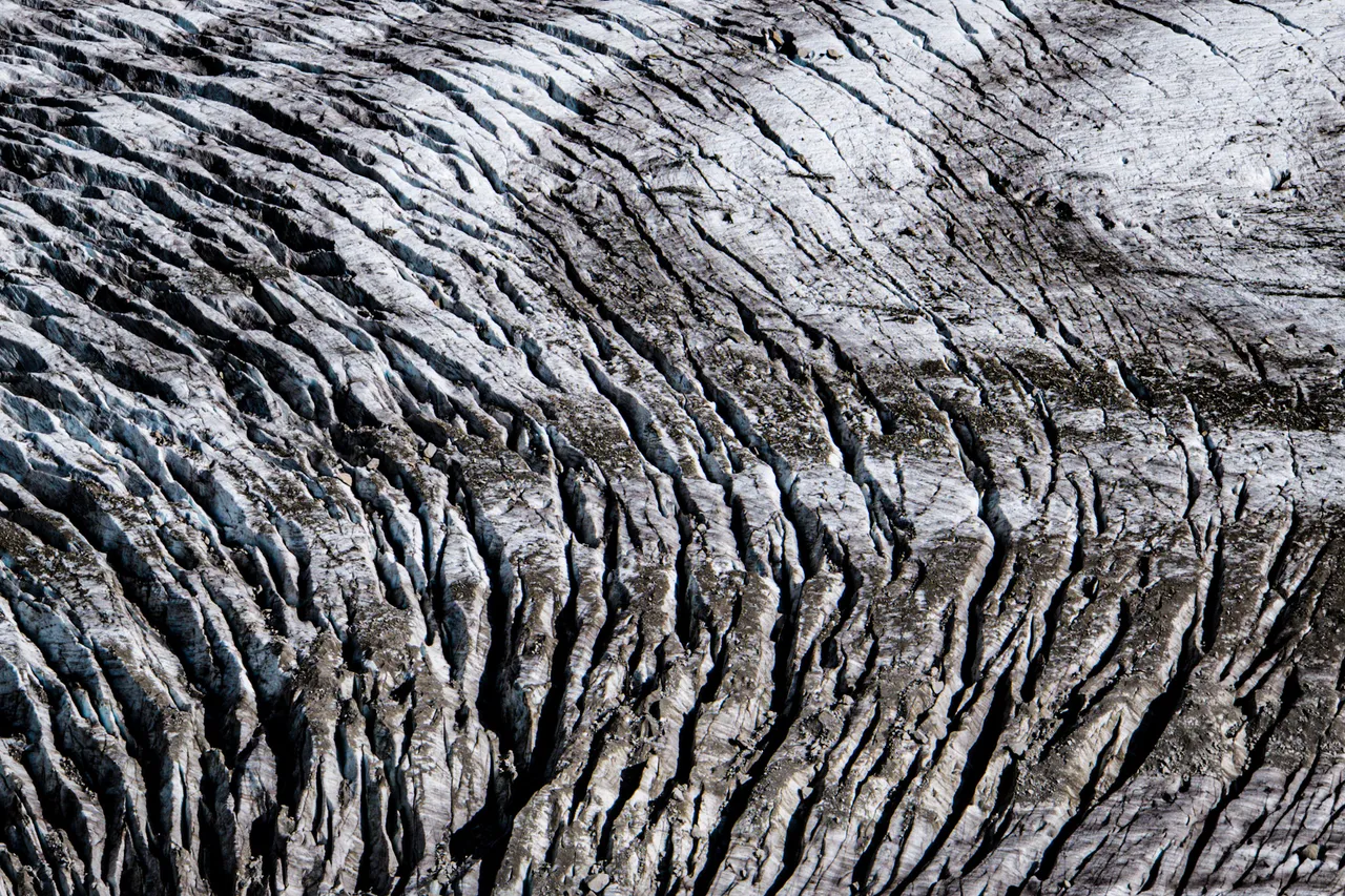 The patterned, cracked surface of a glacier seen from above, in grey and white tones.