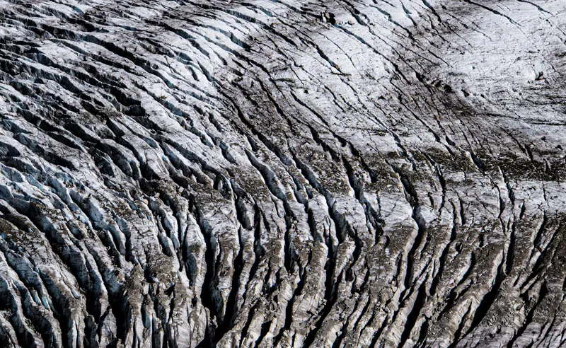 The patterned, cracked surface of a glacier seen from above, in grey and white tones.