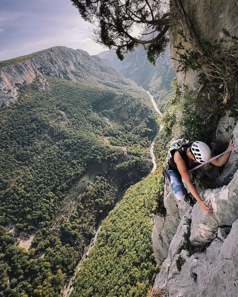 A climber in a white helmet on a pale limestone wall above a deep forested gorge.