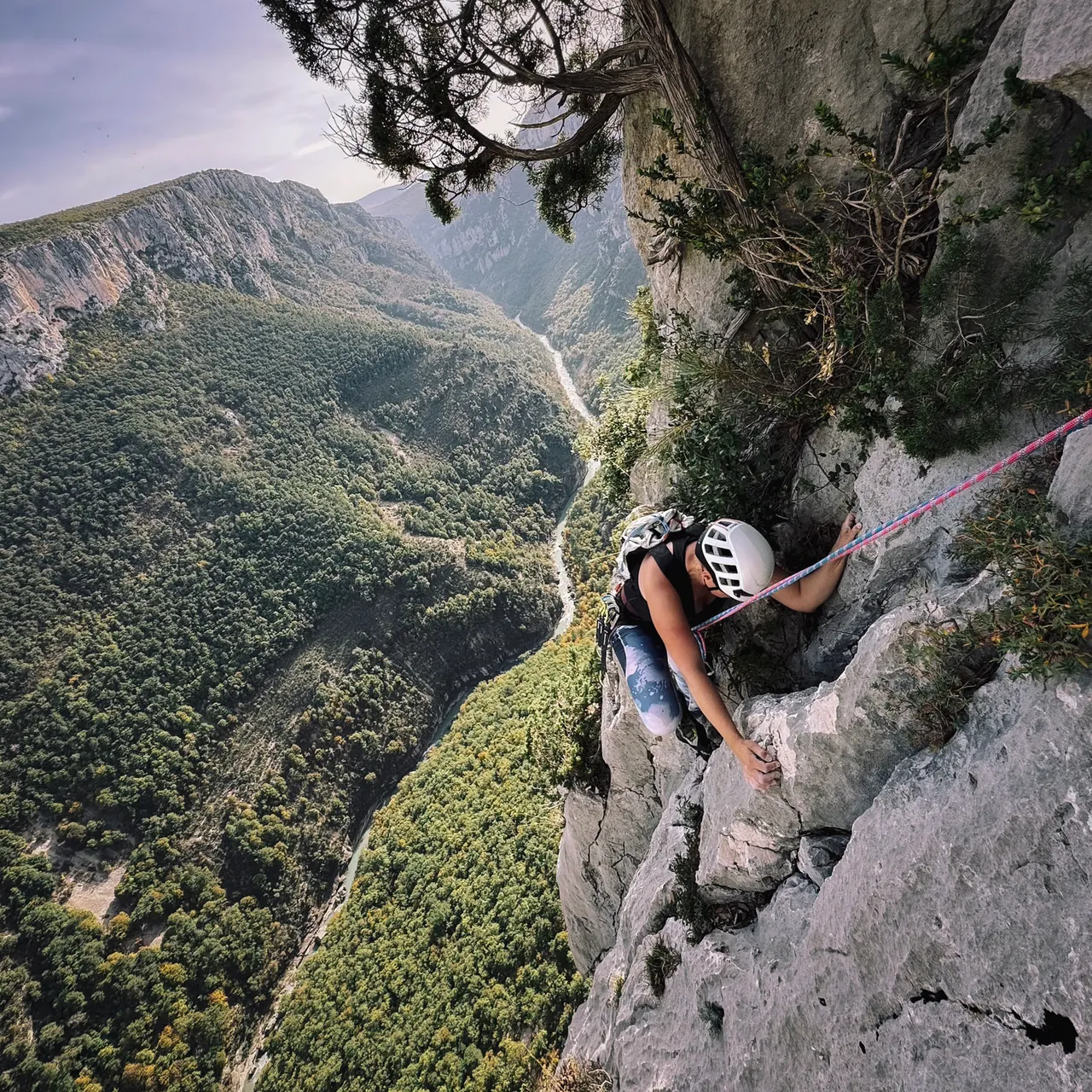 A climber in a white helmet on a pale limestone wall above a deep forested gorge.