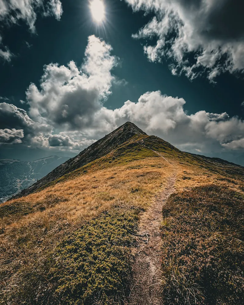 A grassy ridge with a narrow path leading up to a summit, under a sky of big clouds.