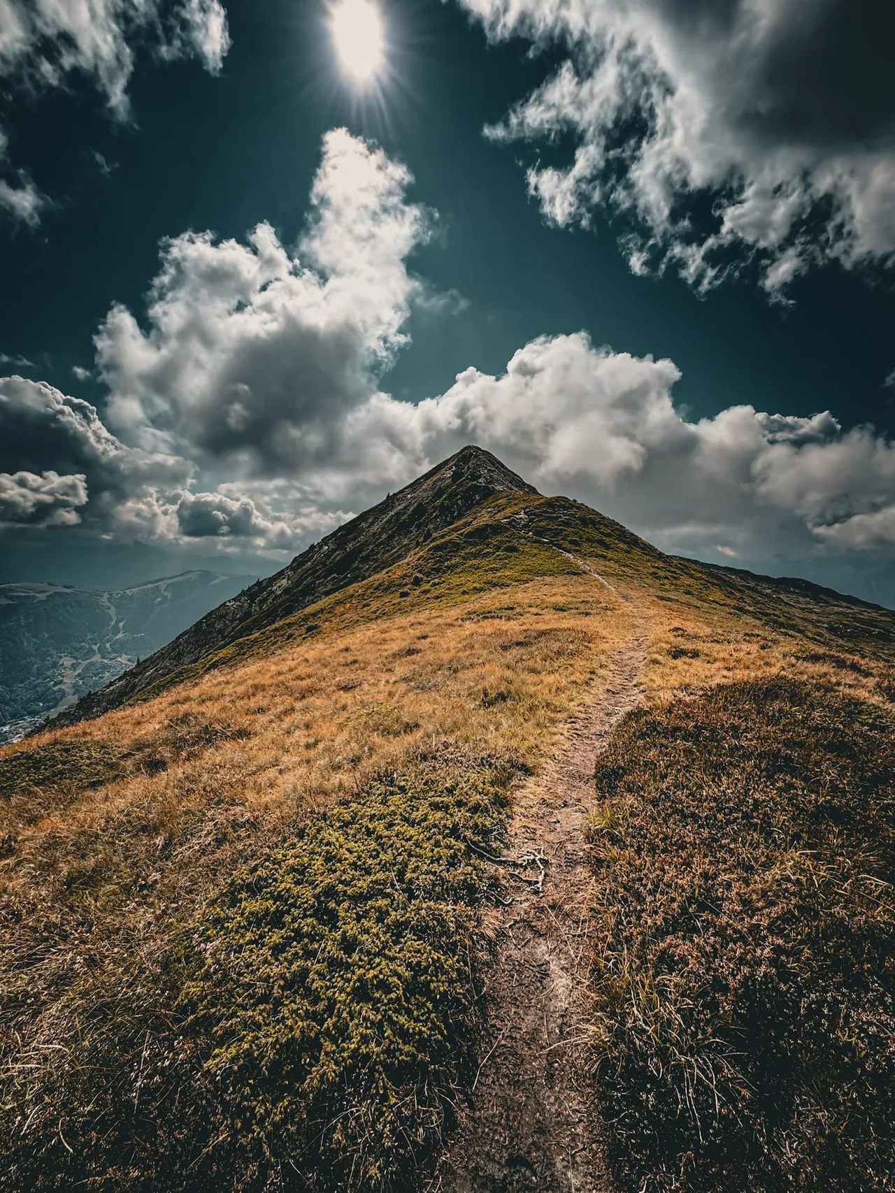 A grassy ridge with a narrow path leading up to a summit, under a sky of big clouds.