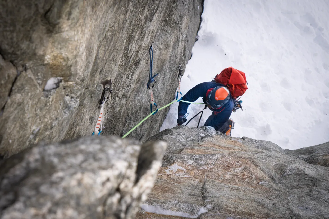 A climber in a red helmet and rucksack moving up a snowy rock step, seen from below.