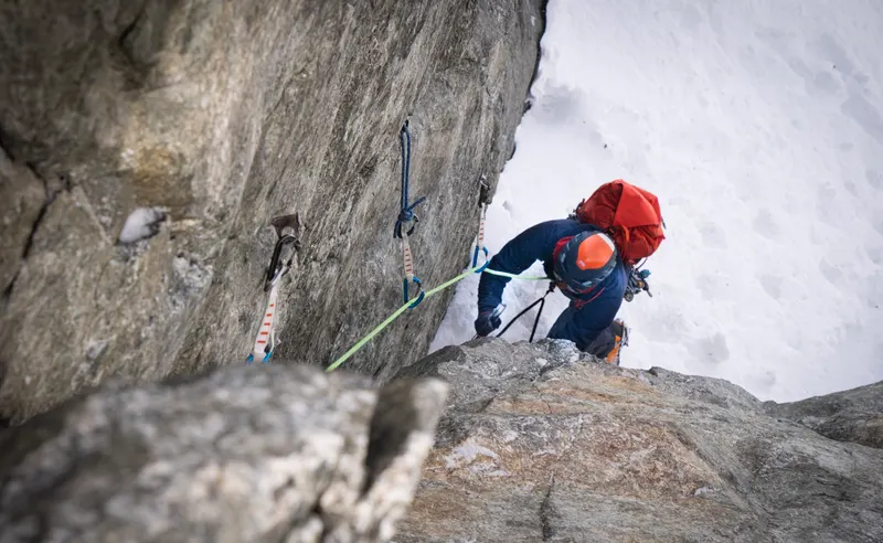A climber in a red helmet and rucksack moving up a snowy rock step, seen from below.