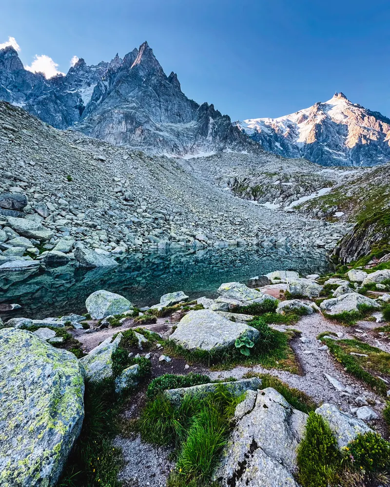 A still alpine lake among granite boulders and scree, below jagged peaks and a snow-covered summit in clear morning light.