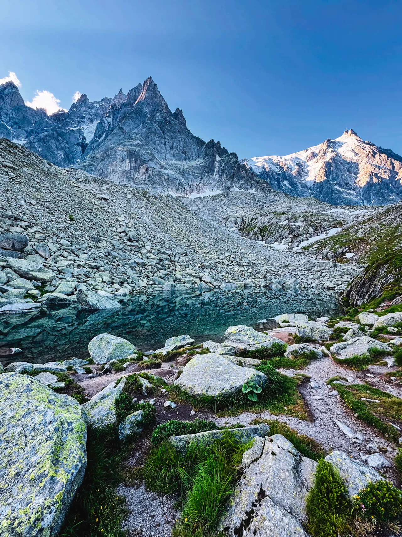A still alpine lake among granite boulders and scree, below jagged peaks and a snow-covered summit in clear morning light.