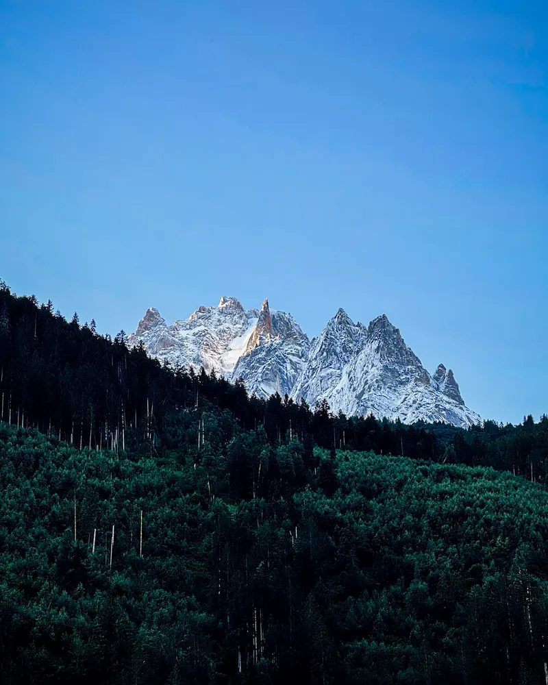 A cluster of snowy granite spires above a dark forested slope, under a clear blue sky.