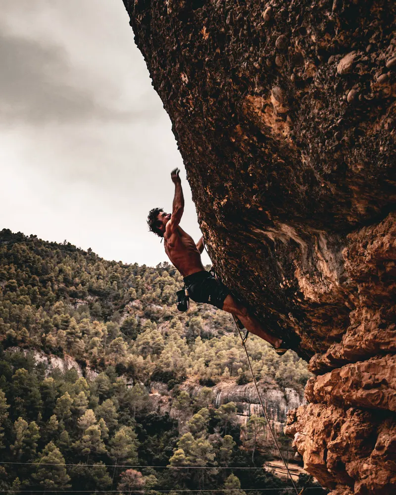 A climber hanging from the underside of a big orange overhang, above a forested hillside.