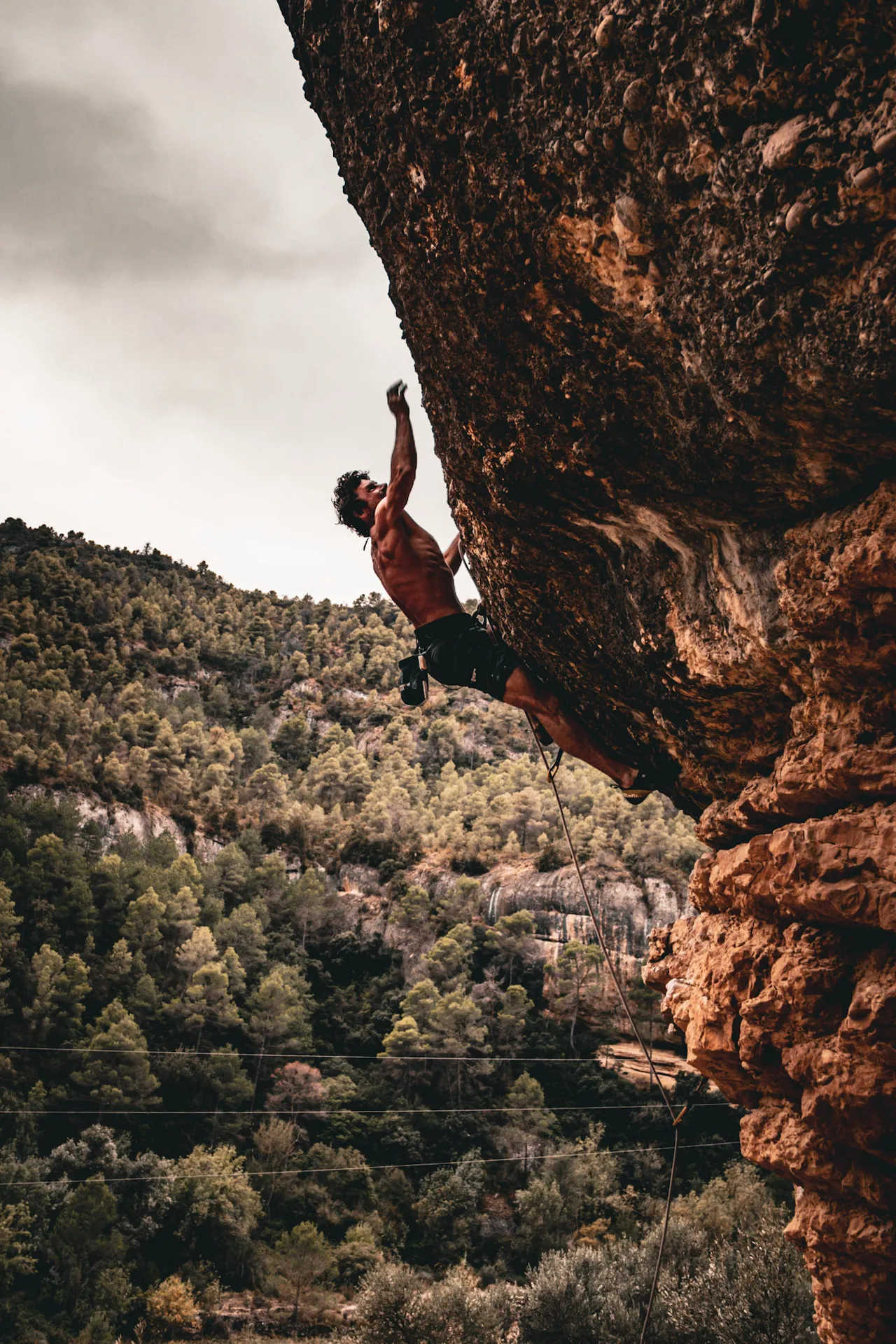 A climber hanging from the underside of a big orange overhang, above a forested hillside.