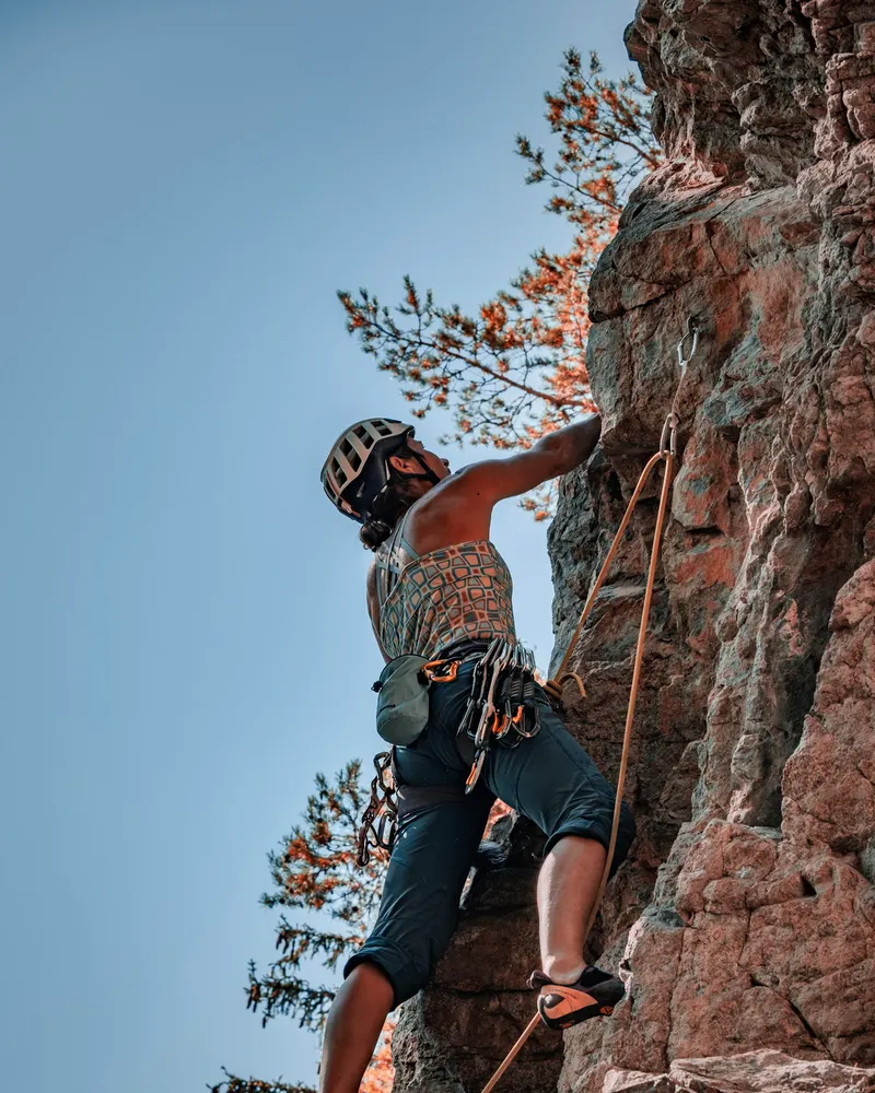 A climber in a helmet on a steep red rock face, with pine branches against a blue sky.