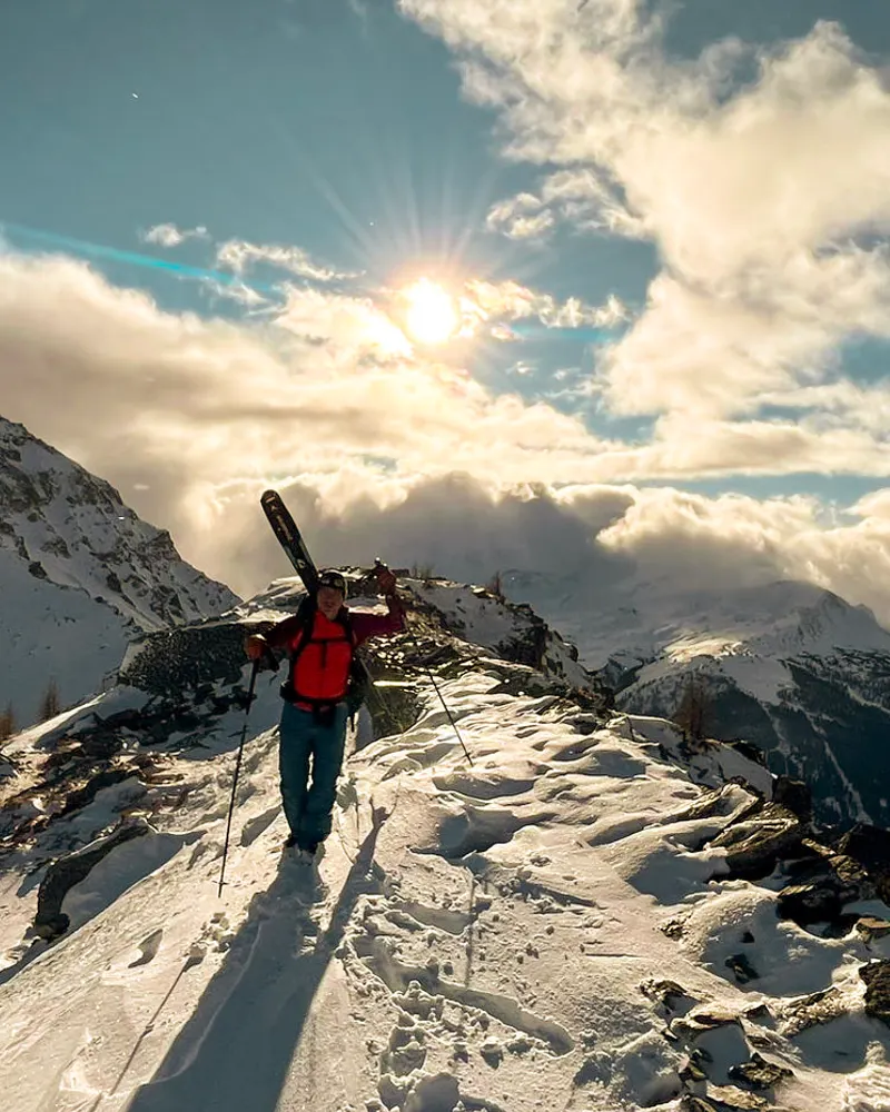 A person with skis on their pack walking a snowy ridge towards a low sun breaking through cloud.