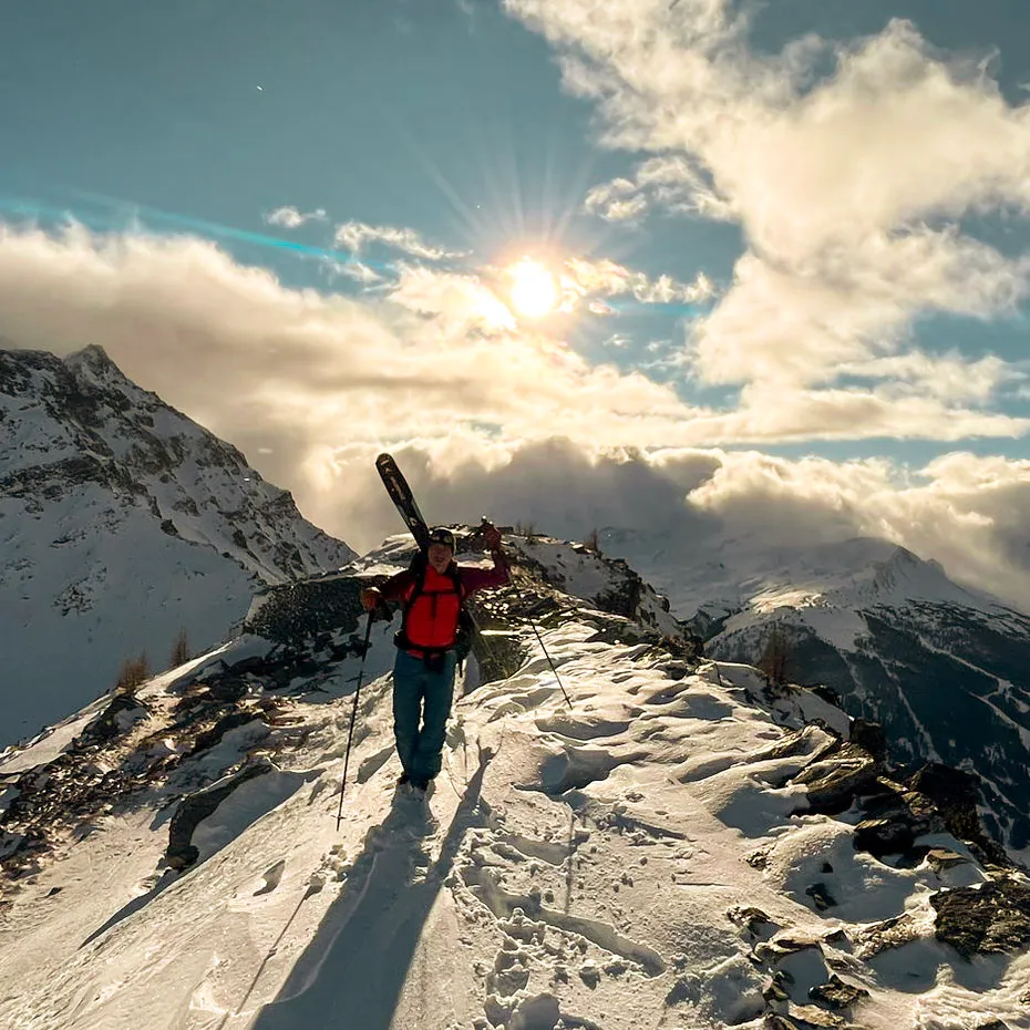 A person with skis on their pack walking a snowy ridge towards a low sun breaking through cloud.