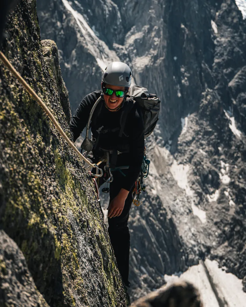 A climber in a helmet and sunglasses on a granite face, tied in to a rope and looking down at the camera.
