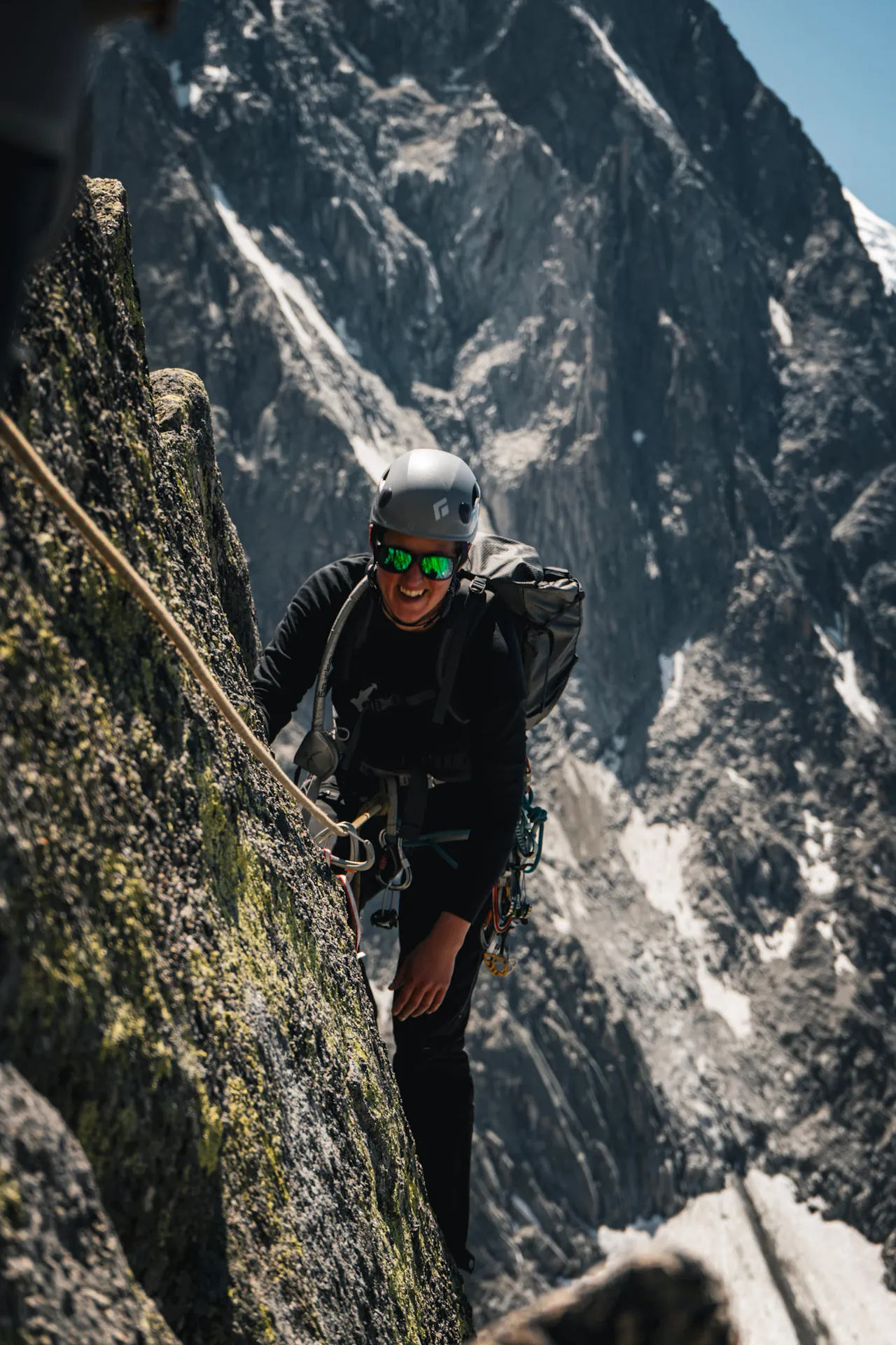 A climber in a helmet and sunglasses on a granite face, tied in to a rope and looking down at the camera.