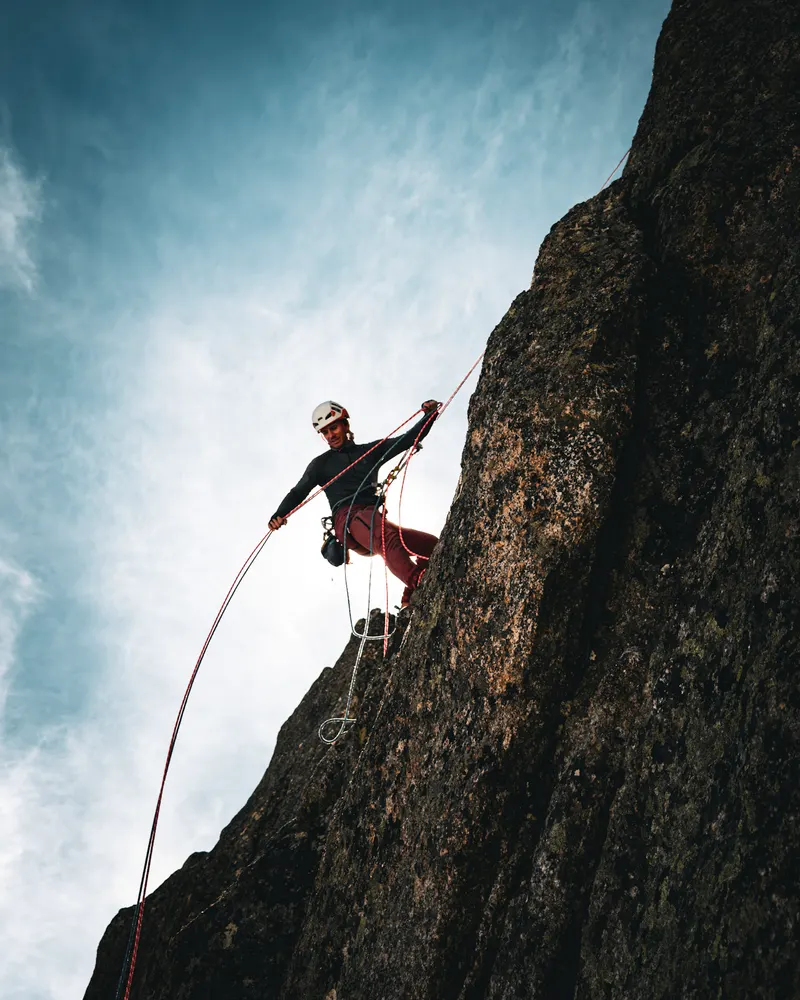 A climber hanging on a rope beside a steep dark rock wall, against a bright sky.