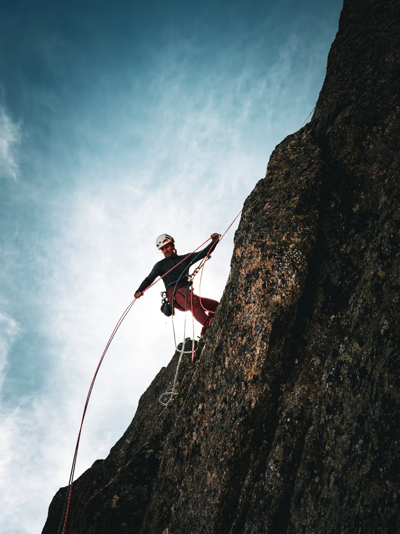 A climber hanging on a rope beside a steep dark rock wall, against a bright sky.