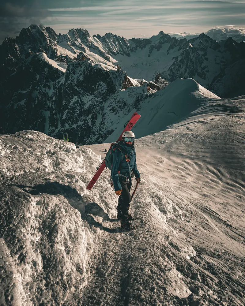 A skier with skis on their pack, walking along a snowy ridge with distant peaks behind.
