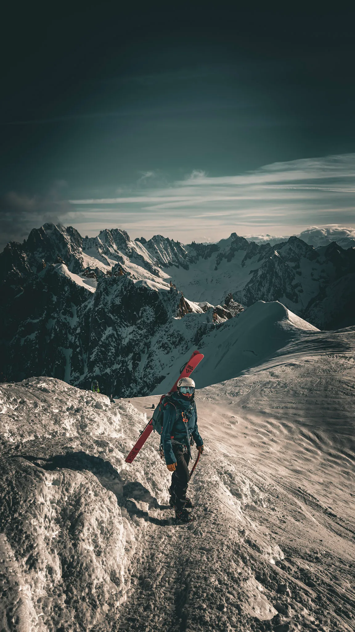 A skier with skis on their pack, walking along a snowy ridge with distant peaks behind.