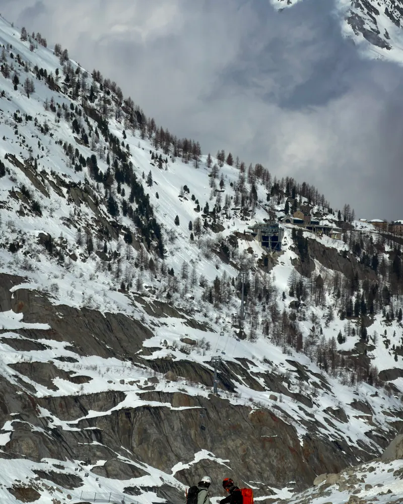 Two small figures on skis on a snowy slope below a rocky mountainside.