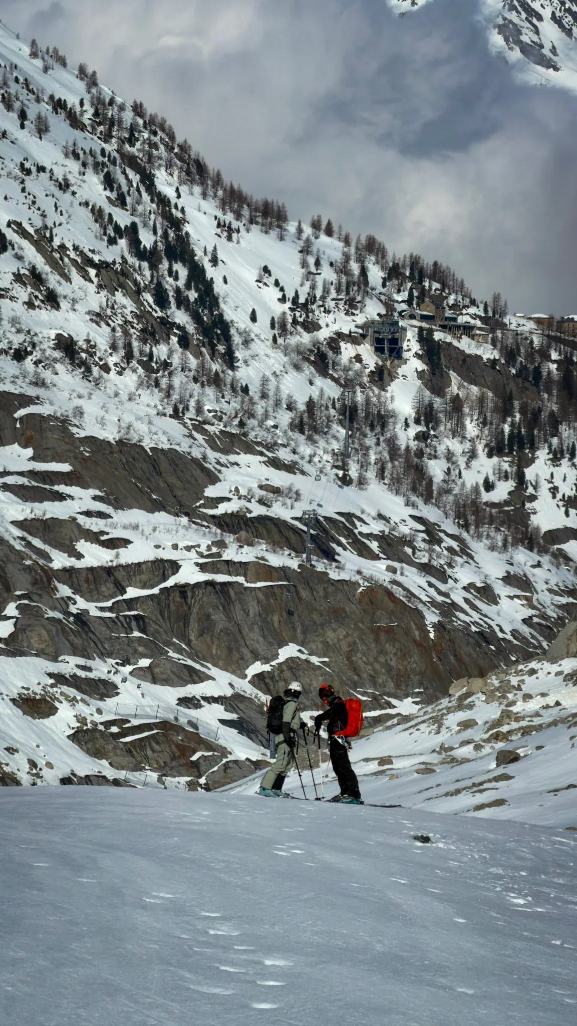 Two small figures on skis on a snowy slope below a rocky mountainside.