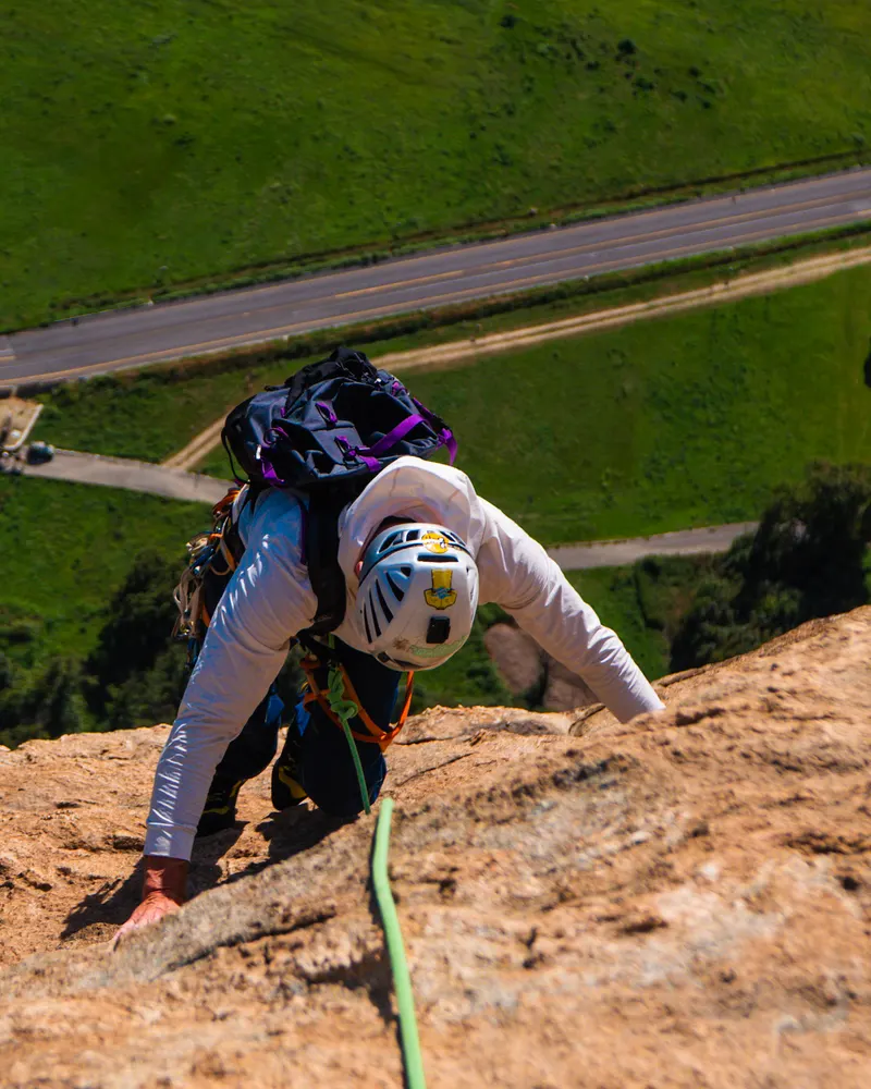 A climber in a white helmet on a warm rock slab, seen from above, with a green valley and a road far below.