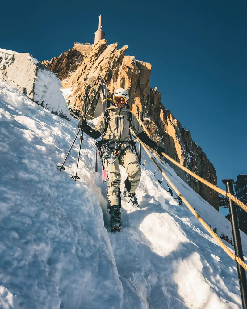 A person with trekking poles walking down a snowy ridge towards a rock tower with a summit station on top.