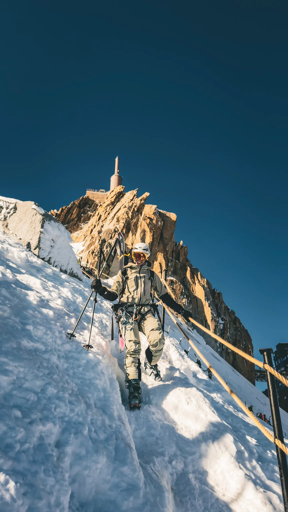 A person with trekking poles walking down a snowy ridge towards a rock tower with a summit station on top.