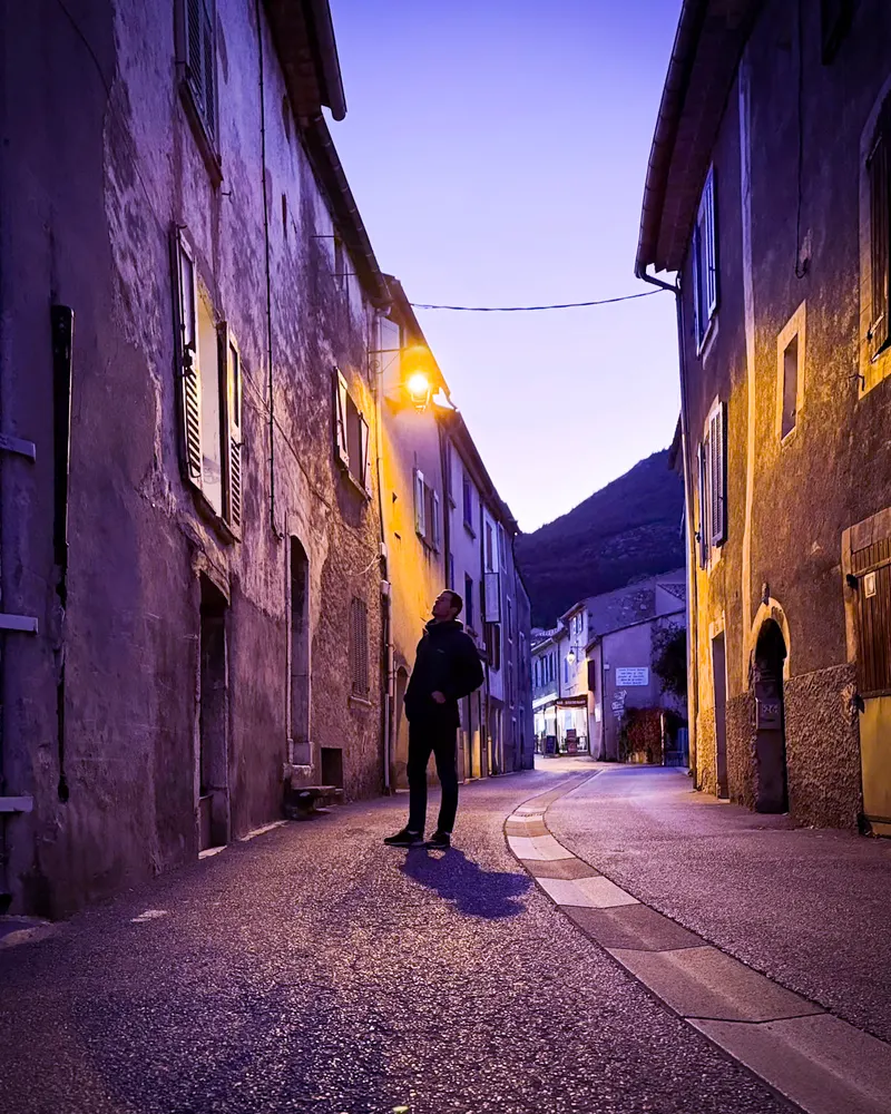 A person walking along a narrow old village street at dusk, with lit windows and a street lamp.