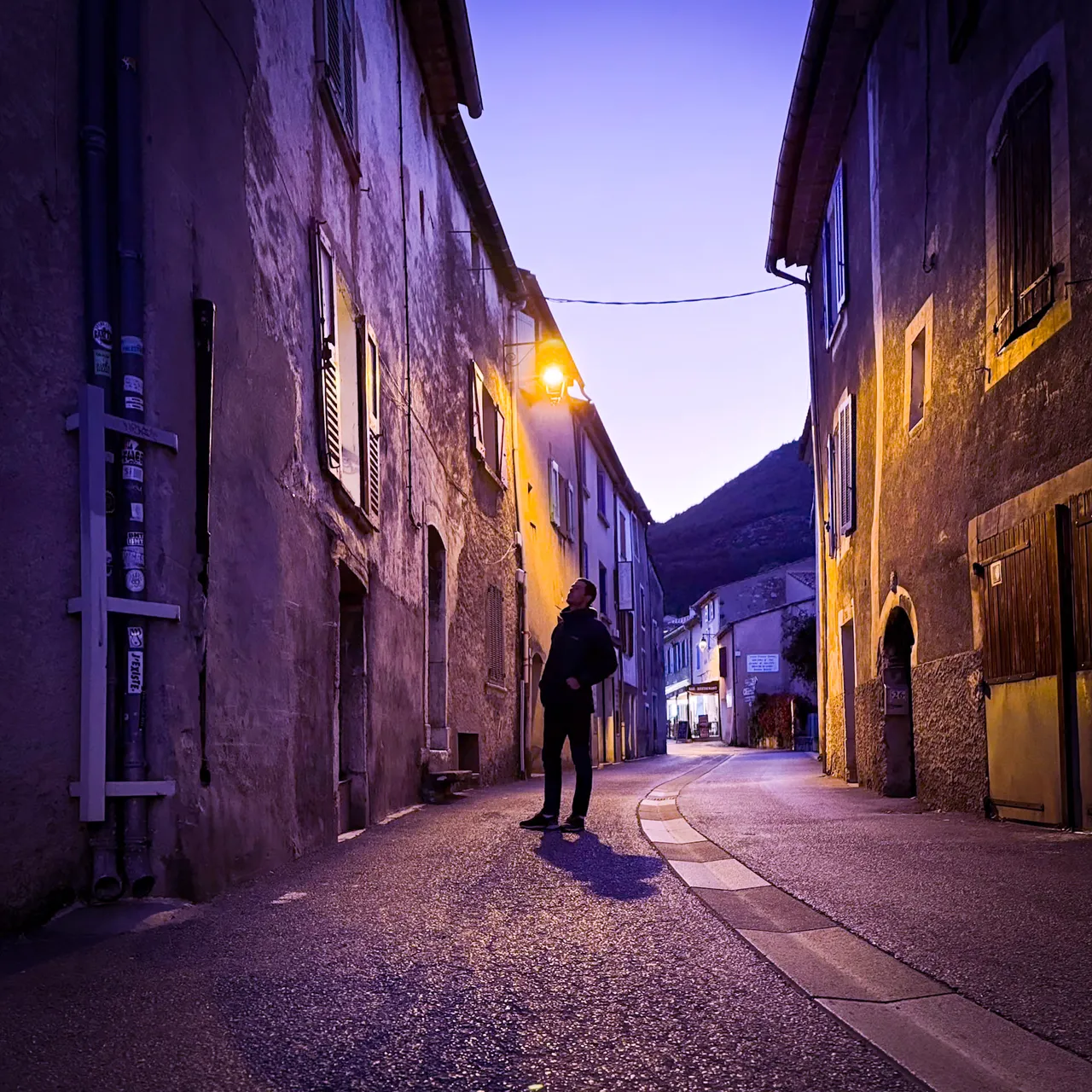 A person walking along a narrow old village street at dusk, with lit windows and a street lamp.