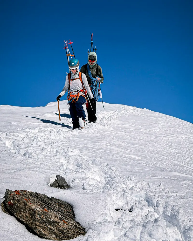 Two people with skis on their packs, climbing a snowy slope under a deep blue sky.