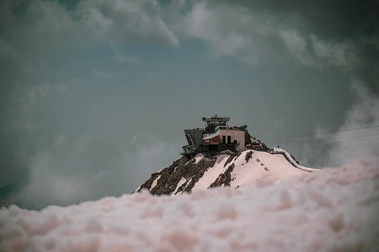 A mountain station on a snowy rock summit, rising above a layer of pink-tinged cloud.
