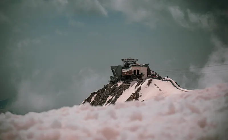 A mountain station on a snowy rock summit, rising above a layer of pink-tinged cloud.