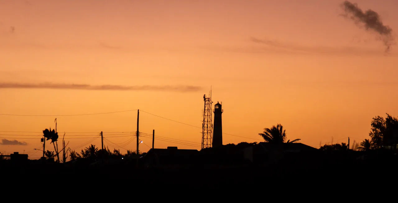 An orange sunset sky over the dark silhouettes of palm trees, a mast and a small tower.