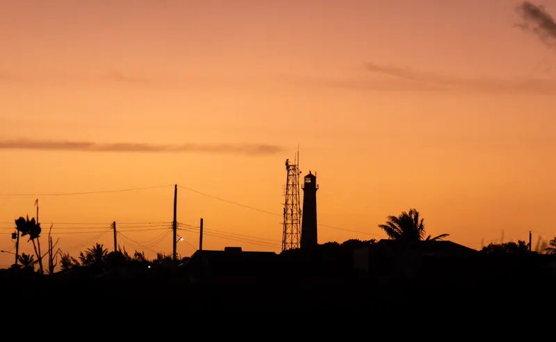 An orange sunset sky over the dark silhouettes of palm trees, a mast and a small tower.