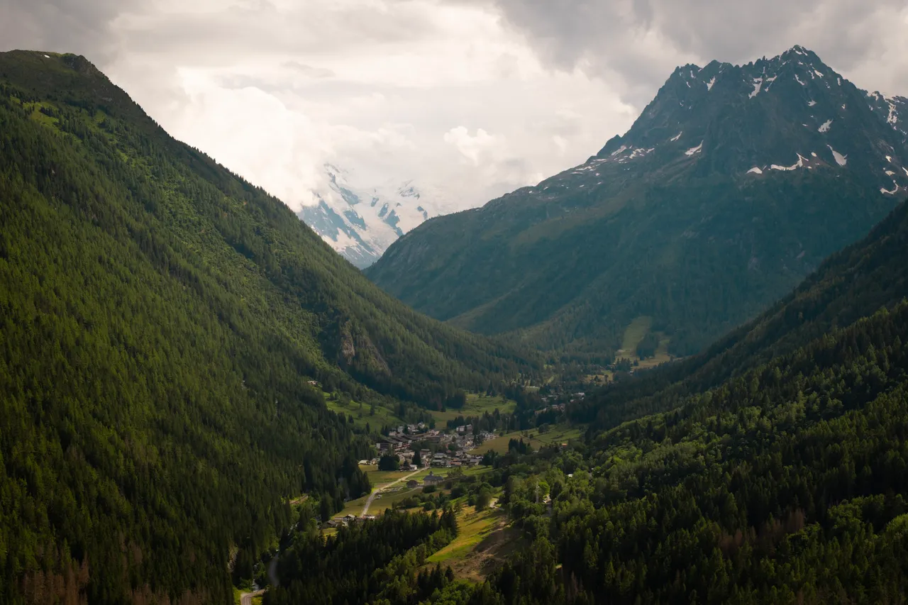 A green valley with a village in the distance, below a snowy peak and a cloudy sky.