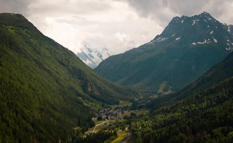 A green valley with a village in the distance, below a snowy peak and a cloudy sky.