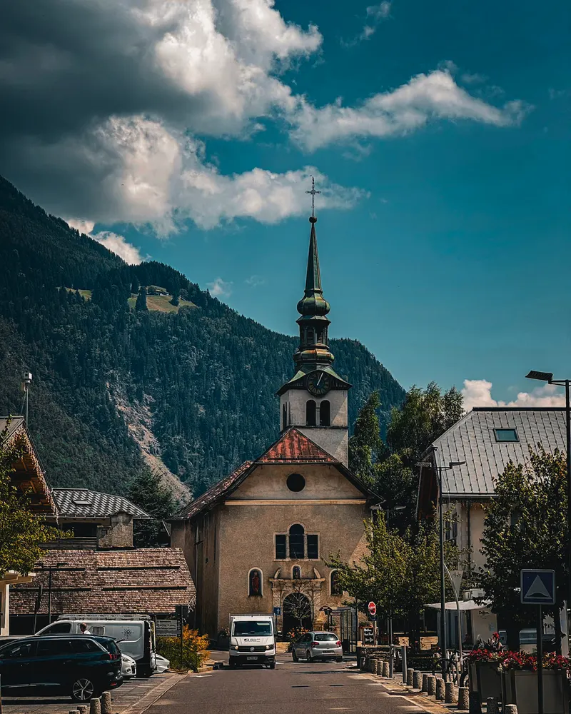 A village street with a stone church and spire, below forested mountains and a blue sky with clouds.