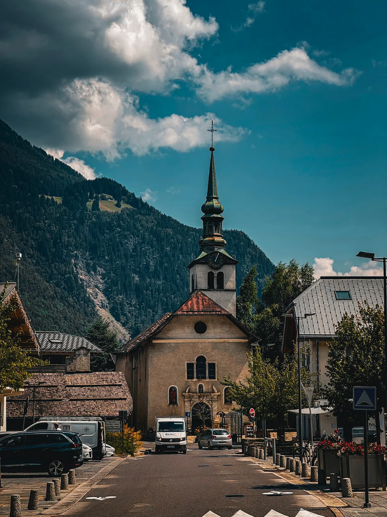 A village street with a stone church and spire, below forested mountains and a blue sky with clouds.